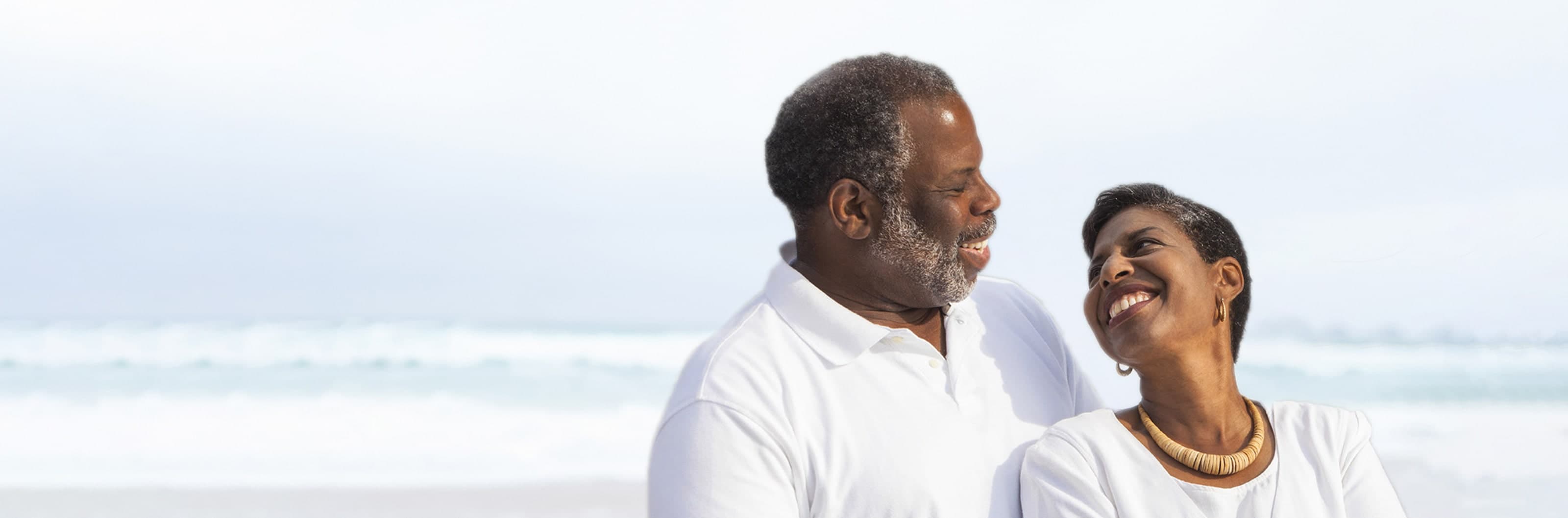 Two older adults standing together on a beach with ocean waves visible in the background.