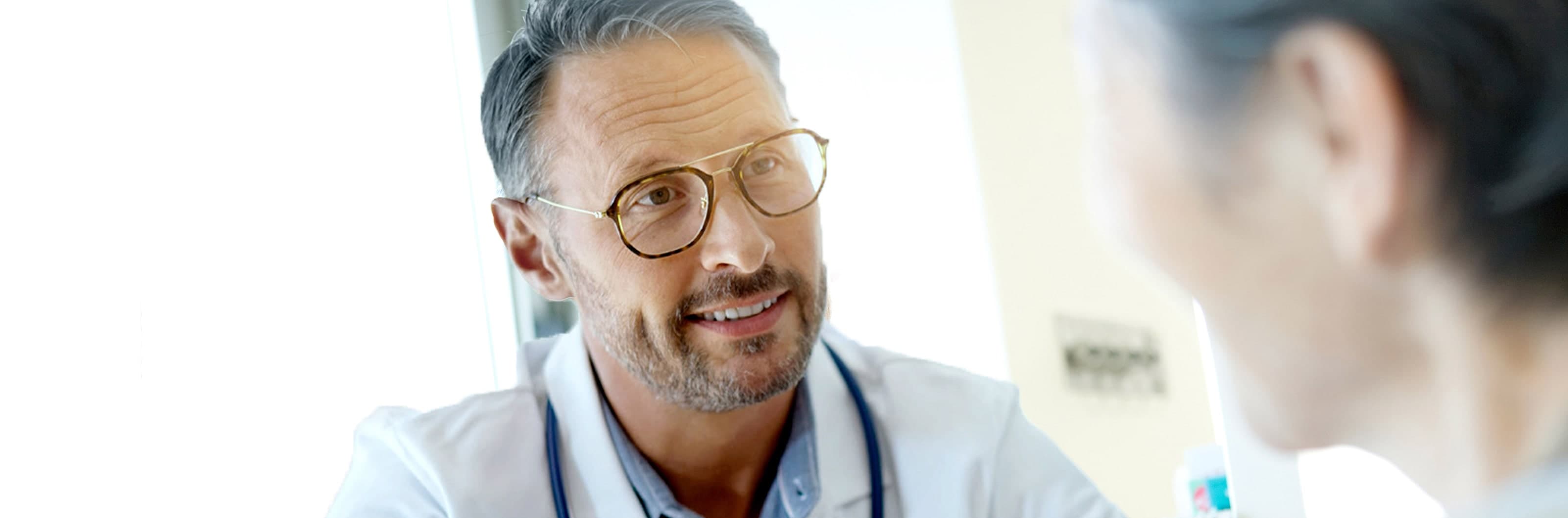 Close-up of a doctor wearing glasses and a lab coat speaking with a patient in a bright clinical setting.